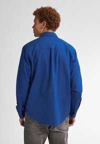 Modrá košile s dlouhým rukávem, zapínáním na knoflíky a zakulaceným spodním lemem. Materiál vypadá hladce, má jedinou zadní yoke a knoflíky na manžetách.