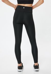 Leggings noirs taille haute avec une texture lisse, un design sans couture et un détail logo sur la ceinture. Portés avec des chaussures de sport noires.