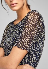 Marinblus med ett genomskinligt, mönstrat tyg som har abstrakta prickar i grön, gul och vit; korta ärmar med scallopade kanter.