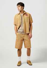 Kortärmad, beige randig skjorta med fickor och en patch, tillsammans med matchande randiga shorts. Har en rundhalsad, beige t-shirt under.