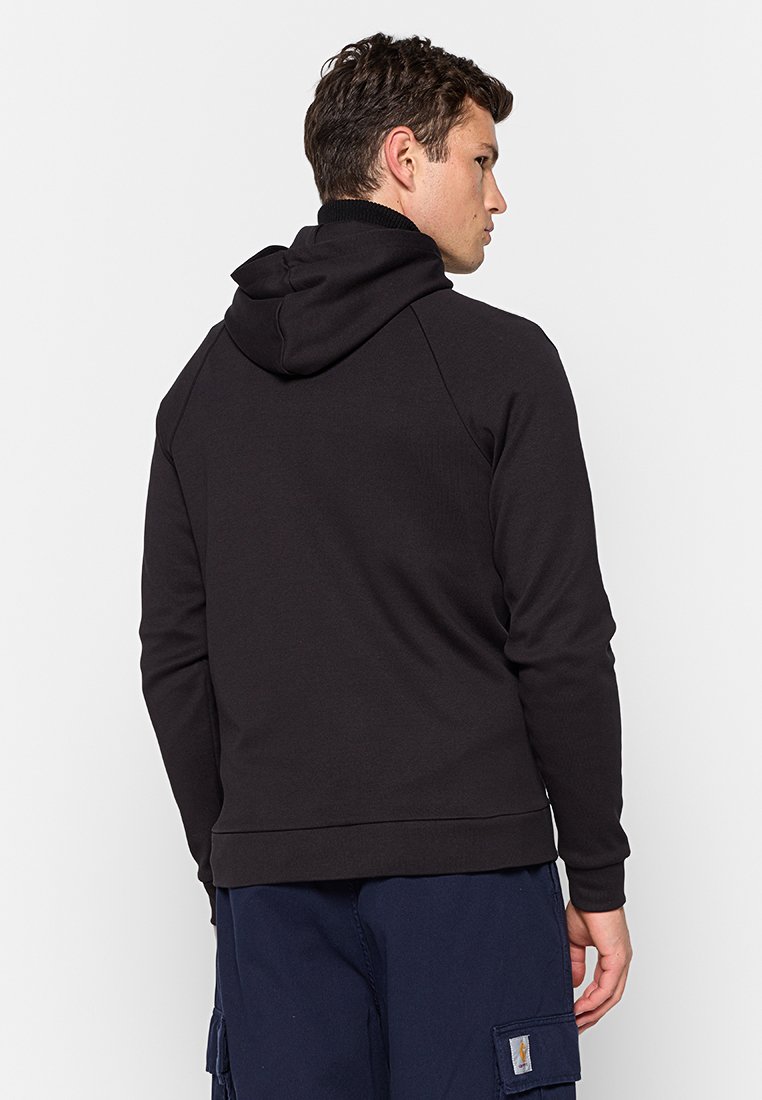 Man met zijn rug naar de camera, gekleed in een zwart hoodie en marineblauwe cargo-broek, tegen een effen witte achtergrond.