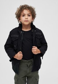 Juodas užtrauktuku užsegamas švarkas su dviem priekiniais kišeniais, dėvimas ant juodo marškinėlio. Derinamas su alyvuogių žaliais cargo kelnėmis, kurios turi šonines kišenes ir laisvą siluetą.