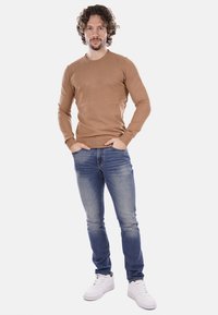 Camicia marrone a maniche lunghe abbinata a jeans azzurri e sneakers bianche. Tessuto morbido con un design aderente e scollo rotondo.