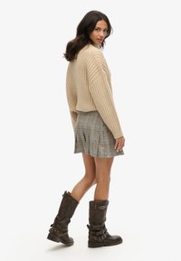 Beige geribbelde cropped trui, geruite geplooide minirok en bruine leren kniehooge laarzen met gespen. Model staat met de rug naar de kijker.