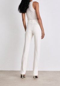 Pantalon blanc sur mesure avec une coupe droite, texture lisse, taille haute et détail de couture discrète à l'arrière, associé à un débardeur simple.