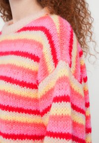 Άτομο με σγουρά μαλλιά που φοράει ένα χνουδωτό ριγέ πουλόβερ σε ροζ, κόκκινο και κίτρινο χρώμα, ορατό από την περιοχή των ώμων και του λαιμού.
