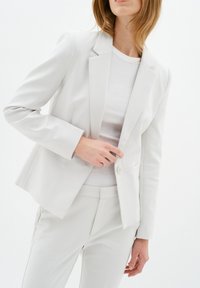 Světlé šedé přizpůsobené sako s jedním knoflíkem a klopovými límci, nošené přes bílé tričko, s elegantním designem a rovný fit.
