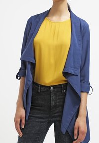 Cardigan ouvert léger bleu marine avec des manches retroussées sur un chemisier jaune. Associé à un jean foncé et ajusté, créant un look superposé décontracté.
