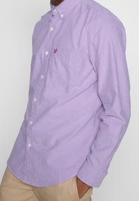 Camicia button-up lilla in cotone, con una piccola tasca sul petto e un accento con logo. Presenta un colletto a punta e maniche lunghe.