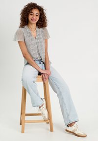 Blouse à manches courtes, à motifs, de couleur claire, associée à un jean large léger bleu. Le modèle est assis sur un tabouret en bois portant des baskets.
