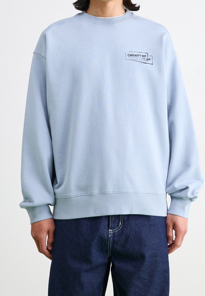 Sudadera azul claro hecha de un tejido suave, con cuello redondo y puños acanalados. Un pequeño parche de logo negro está en el pecho.