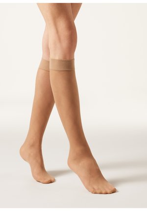 Beige knähöga strumpor i genomskinligt material med en ribbad toppkant och subtilt diamantmönster längs benen, som ger en slät textur.