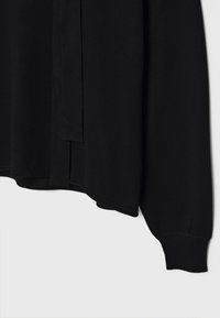 Haut noir en maille à manches longues avec un poignet côtelé et un large détail noué en tissu qui pend à l'avant.