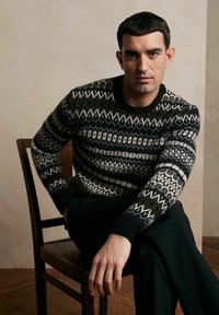 Sweter dzianinowy z czarno-kremowym geometrycznym wzorem, okrągłym dekoltem i ściągaczami na rękawach. Model siedzący na drewnianym krześle, w czarnych spodniach.