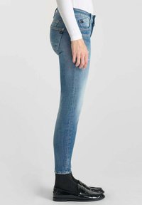 Jeans skinny en denim bleu clair, présentant un délavage, des poches arrière et une taille mi-haute, associés à des mocassins noirs en cuir verni et des chaussettes.