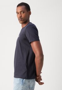 Tamno plavi pamučni T-shirt s kratkim rukavima, okruglim izrezom i suptilnim logotipom na prsima. U kombinaciji s svijetloplavim trapericama.
