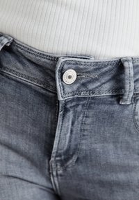 Svijetloplavi traperici s glatkom teksturom, koji imaju kopčanje na dugme i blago izblijeđeli finiš. Pojas je uparen s rebrastim bijelim topom.