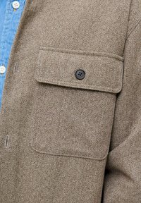 Brun tekstureret skjortejakke med en klaplomme og en sort knap, lagdelt over en lyseblå denimskjorte med hvide knapper.