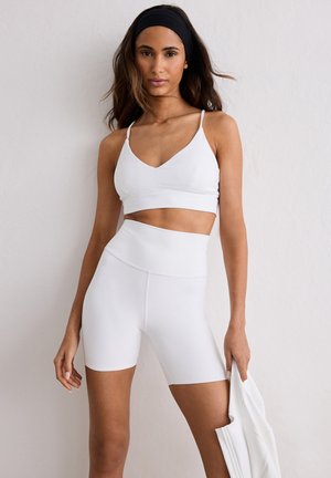 Kvinde iført hvid sports-bh og cykelshorts med høj talje, der holder et hvidt stykke tøj og står foran en lys væg.