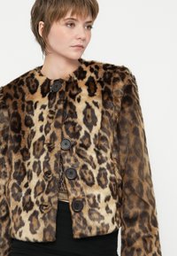 Rövid hajú nő barna és fekete leopárdmintás műszőrme kabátot visel nagy fekete gombokkal egy fekete felső fölött, jobbra nézve.