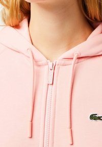 Rosa zip-upp-hoodie i mjukt tyg, med dragsko och en liten grön Krokodil-logotyp på vänster bröst.