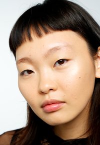 Primo piano di un volto con pelle chiara, leggere imperfezioni, capelli scuri e lisci con frangia dritta e colore delle labbra neutro. Lo sfondo è bianco uniforme.
