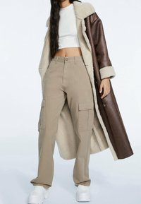 Hnědo-beige kabát z beránka přes bílý crop top, doplněný béžovými cargo kalhotami. Bílé tenisky outfit dokončují.