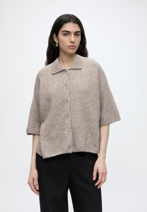 Kvinde med langt mørkt hår iført en beige kortærmet knap-op sweater og sorte bukser, stående mod en ensfarvet lys baggrund.