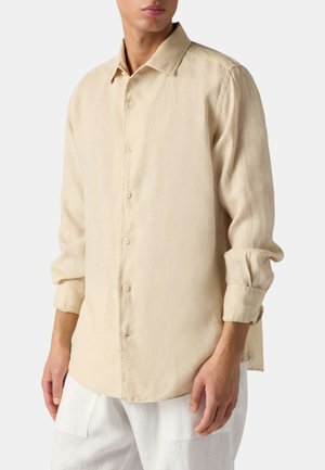 Mand iført en beige langærmet skjorte med knapper og opsmøgede ærmer samt hvide bukser mod en ensfarvet hvid baggrund.