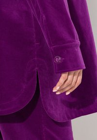 Giacca in velluto a coste viola con polsini abbottonati, che presenta linee verticali testurizzate e una fessura laterale per un tocco di dettaglio in più.