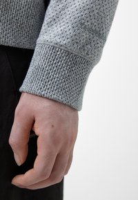 Grå teksturert genserarm har et pustende, perforert design med en ribbet mansjett. Subtil merkevarebygging er synlig på materialet.