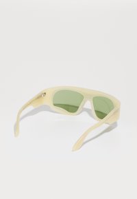Cream-colored sunglasses with tinted green lenses, a sleek design, and subtle branding on the side. Smooth texture with rounded edges.