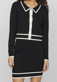 Svart stickad klänning med långa ärmar, vita detaljer och krage. Har en knäppning framtill och horisontella ränder vid midjan och nederkanten.