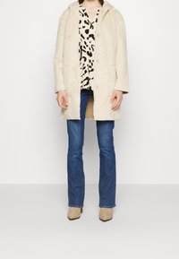 Personne portant un manteau duveteux couleur crème sur un chemisier blanc à pois noirs, un jean bleu et des bottines beiges, debout devant un fond uni.