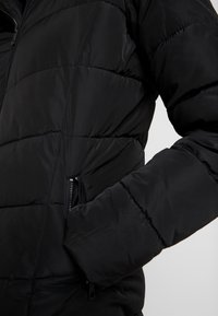 Doudoune noire rembourrée avec des coutures horizontales, un col montant et une poche zippée sur le côté. Le matériau semble lisse et isolant.