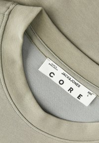 Κοντινή λήψη στο γιακά ενός μπεζ T-shirt Jacks & Jones CORE με στρογγυλή λαιμόκοψη και ετικέτα μεγέθους L με την ένδειξη «Made in Bangladesh.»