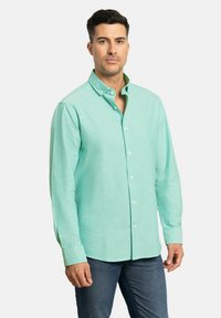 Camisa de botones verde claro con mangas largas, fabricada en un tejido texturizado y con cuello de botón, combinada con vaqueros oscuros.