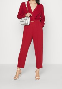 Mono rojo con escote en V profundo, mangas largas y pantalones ajustados. Combinado con un bolso blanco que presenta un patrón sutil y herrajes dorados.