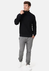 Maglione nero a collo alto in maglia con texture a coste, indossato con pantaloni grigi e sneakers bianche, caratterizzato da una vestibilità comoda e maniche inserite.