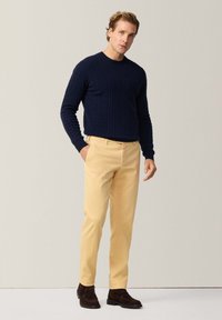 Navy gebreide trui met een textuurpatroon, gecombineerd met lichtgele broek en donkerbruine suède schoenen. Staande pose met de hand in de zak.