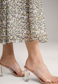 Transparente Blockabsatz-Mules mit einem klaren Riemen, beigem Innensohle und eckigem transparentem Blockabsatz, präsentiert vor einem floral gemusterten Hintergrund.
