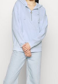 Sudadera con cremallera de color azul claro en tejido suave, con un bolsillo frontal, capucha con cordones y puños de canalé. Combinada con jeans de color azul claro.