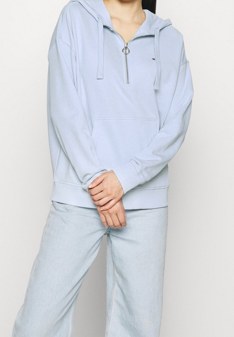 Sudadera con cremallera de color azul claro en tejido suave, con un bolsillo frontal, capucha con cordones y puños de canalé. Combinada con jeans de color azul claro.