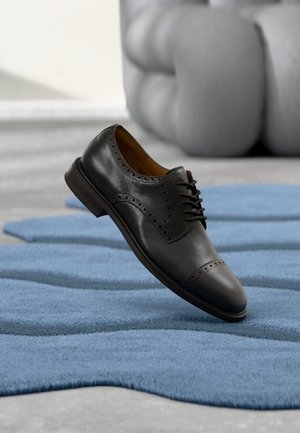 Black leather dress shoe with brogue detailing, rounded toe, and plain laces, hovering above a blue textured rug in a modern setting.