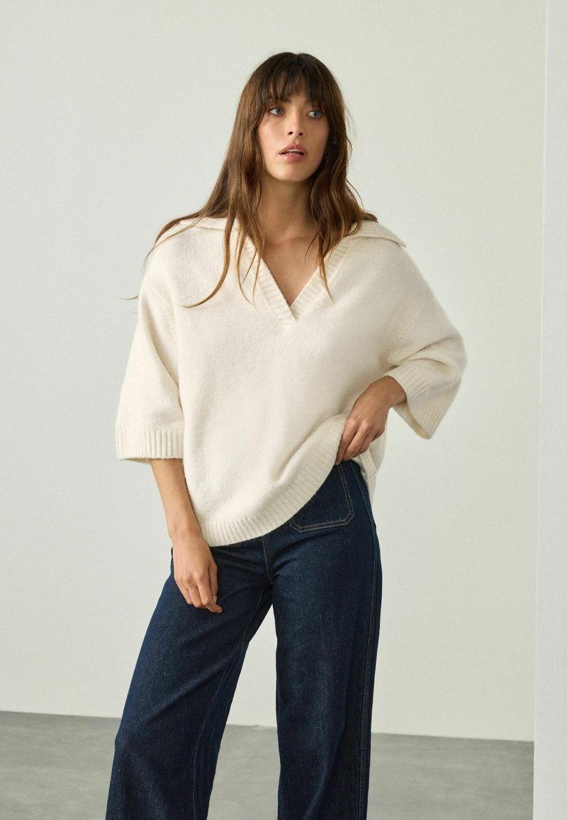 Un pull surdimensionné de couleur crème en tricot doux avec un col en V et des poignets côtelés, associé à un jean large bleu foncé.