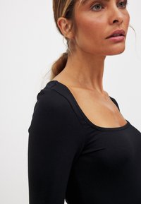 Top nero a maniche lunghe con scollatura ampia, design aderente e texture liscia. Presenta un sottile dettaglio cucito sulla spalla.