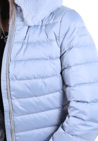 Chaqueta acolchada azul claro con cuello de piel sintética, presenta un diseño acolchado y una cremallera visible. Logo en la manga. Diseño suave y aislante.