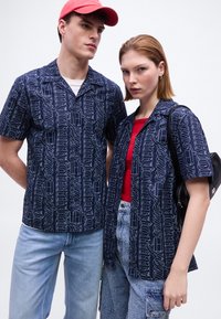 Νέος άνδρας και γυναίκα φορώντας ταιριαστά μπλε πουκάμισα με λευκά γράμματα· ο άνδρας με κόκκινο καπέλο και τζιν, η γυναίκα με κόκκινο τοπ και τζιν σορτς, κρατώντας μαύρο σακίδιο.