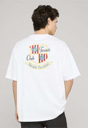 Mand iført hvid oversize T-shirt med rød og gulstribet Tennis Club-våbenskjold og teksten "Sergio Tacchini" på ryggen.