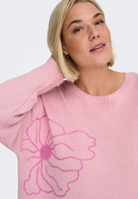 Suéter rosa de punto con un gran diseño de flor en rosa más oscuro en una manga. El escote es de canalé y la textura parece suave.